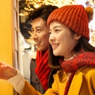 A young couple staring in the window of a store surrounded by Christmas lights and décor.