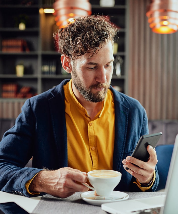 A man holding a cup of coffee looks at his phone for signs of identity theft to avoid.